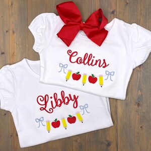 Op de afbeelding: Twee witte T-shirts met rode geborduurde namen "Libby" en "Collins" en een rode strik. Elk shirt heeft een guirlande van rode appels en gele potloden.