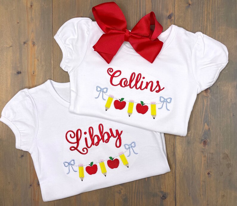 Op de afbeelding: Twee witte T-shirts met rode geborduurde namen "Libby" en "Collins" en een rode strik. Elk shirt heeft een guirlande van rode appels en gele potloden.