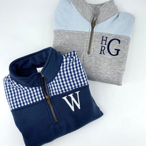 May include: Two quarter-zip sweatshirts with contrasting fabric panels. One is navy blue with white gingham and has a white "W" monogram. The other is gray with a blue and white striped panel and has a blue "HRG" monogram.