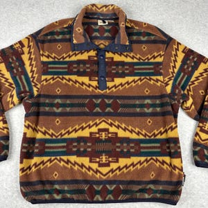 Puede incluir: Un jersey polar marrón con un estampado geométrico en amarillo, verde azulado y granate. El cuello y la tapeta son de color azul marino, con tres botones azul marino. La camisa de manga larga tiene un corte holgado.