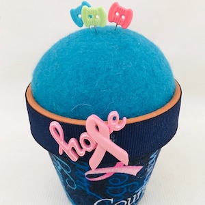 May include: A blue felt pincushion in the shape of a flower pot with a navy blue ribbon and a pink ribbon with the word "hope" on it. The pincushion has three colorful plastic pins on top.