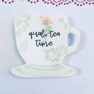 May include: White ceramic teacup and saucer with green and white floral design. The text "quali-tea time" is written on the cup. Two colorful pins are stuck in the cup.