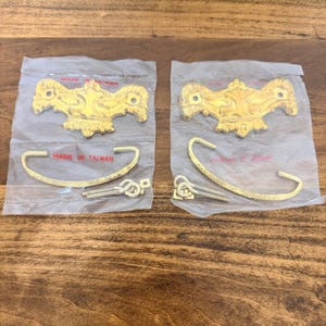 May include: Two sets of gold-toned cabinet hardware, each packaged in clear plastic. Each set includes a decorative handle and a curved pull, along with small connecting hardware. The packaging reads "MADE IN TAIWAN".