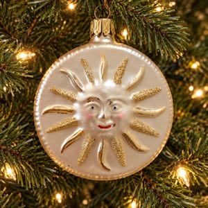 May include: A round, cream-colored glass Christmas ornament featuring a raised sun face with a smiling expression, pink cheeks, and blue eyes. The sun rays are accented with gold glitter, and the ornament has a metallic gold cap at the top.