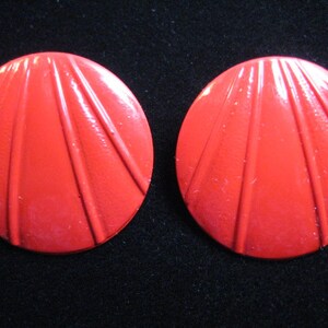 Pode incluir: Um par de brincos redondos de plástico vermelho brilhante com um design de raios de sol.
