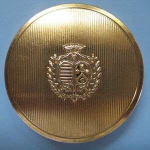 May include: A gold-colored round compact with a raised, detailed crest in the center. The crest features a crown, a shield with a design, and a wreath of leaves.