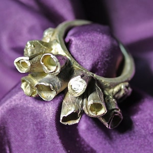 May include: A silver-toned ring with a cluster of tubular, textured elements. The ring is set against a backdrop of a deep purple, silky fabric, creating a contrast in textures and colors. The ring's design has a natural, organic feel.