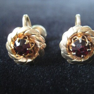 May include: A pair of gold-tone clip-on earrings with a red gemstone in the centre of each earring. The earrings are shaped like a flower.