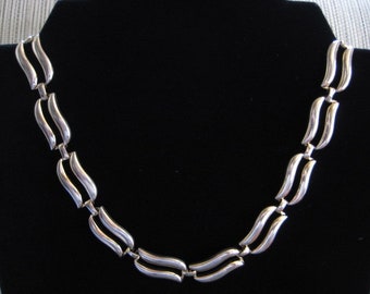 May include: A silver necklace with a wavy, linked chain design. The necklace is displayed on a black background.