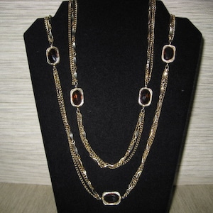 May include: A gold multi-strand necklace with brown oval-shaped stones. The necklace has a layered look with three different chain styles.