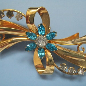 May include: A gold-coloured brooch in a bow design, with a central flower adorned with blue and clear crystal accents. Clear crystals are set along the bow's curves. The brooch has a vintage aesthetic.