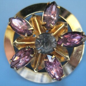 May include: A gold-toned brooch with a circular design featuring a central gray stone surrounded by six purple rhinestones.