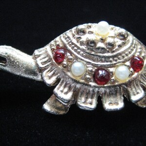 May include: A gold-toned metal turtle brooch with red and white gemstones. The brooch features a detailed shell design with a textured finish. The turtle's head and flippers are also visible, creating a charming accessory.