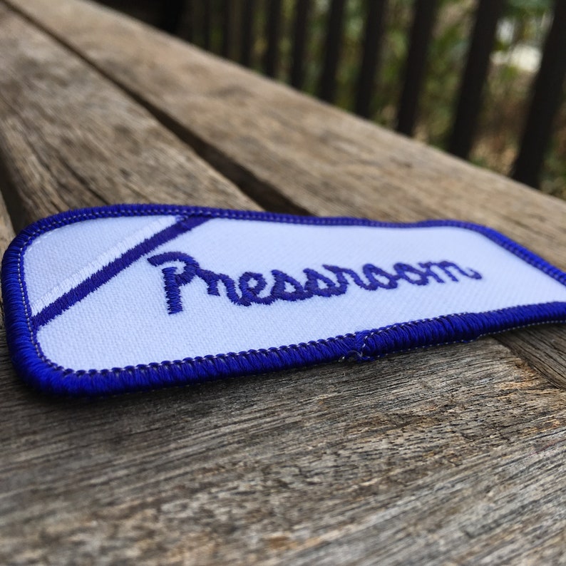 Pressroom A white work shirt uniform patch that says Pressroom in blue script with blue border.