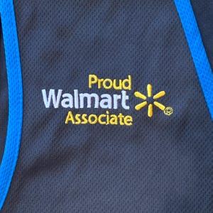 May include: A black vest with a blue trim. The vest has a yellow embroidered logo that says 'Proud Walmart Associate' and a yellow star with a circle around it.