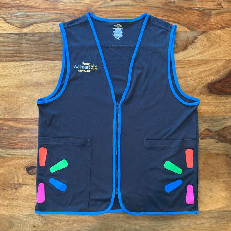 May include: A black vest with blue trim and a zipper. The vest has two pockets on the front and a logo that says 'Proud Walmart Associate' with a yellow star. The vest also has a Walmart logo with a rainbow design on each side.