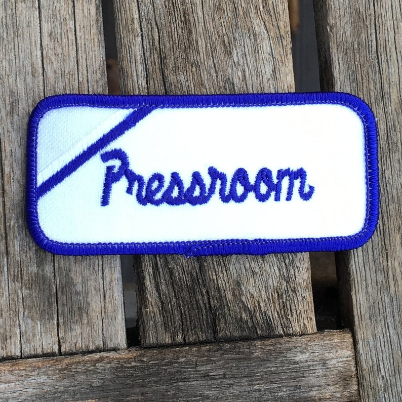 Pressroom A white work shirt uniform patch that says Pressroom in blue script with blue border.