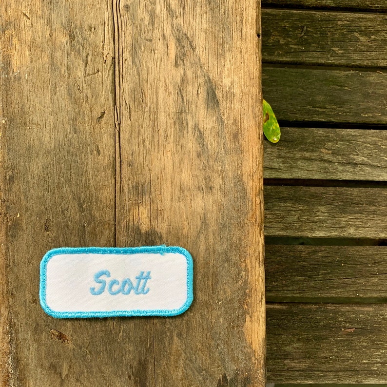 A white work shirt patch that says Scott in light blue script with light blue border. Scott