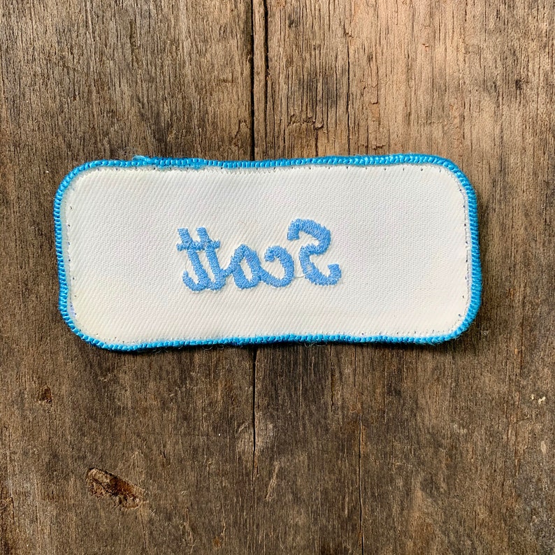 A white work shirt patch that says Scott in light blue script with light blue border. Scott