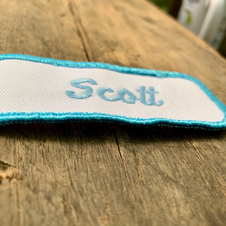 A white work shirt patch that says Scott in light blue script with light blue border. Scott
