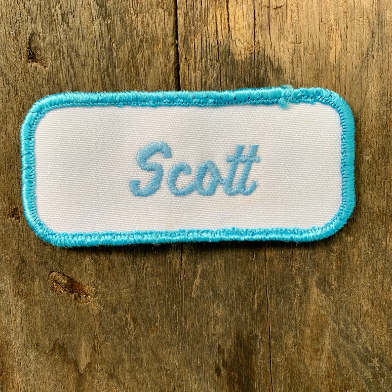 A white work shirt patch that says Scott in light blue script with light blue border. Scott