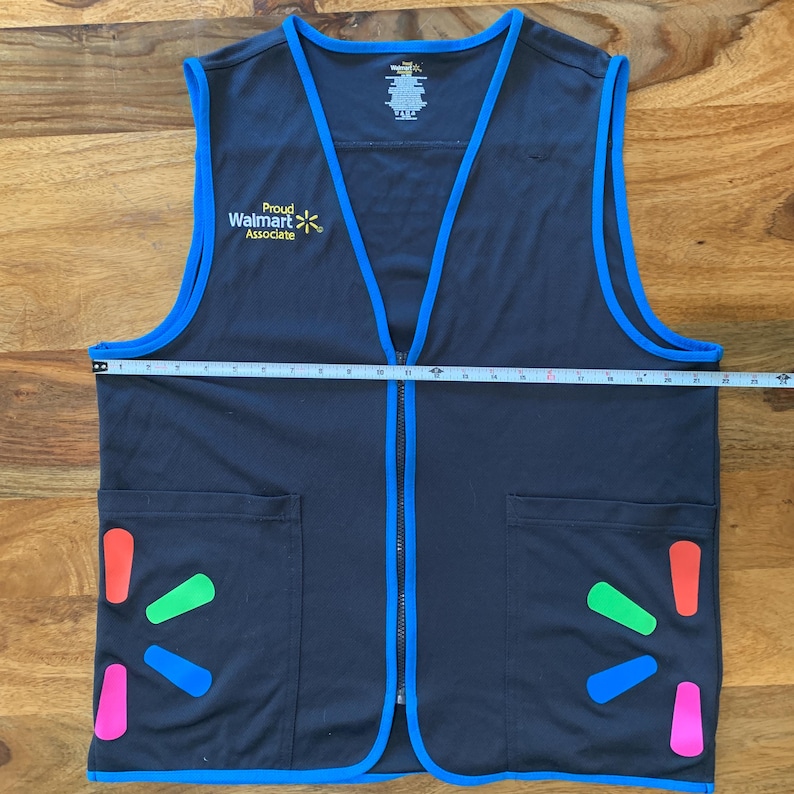 May include: A black vest with blue trim and a zipper closure. The vest has two pockets on the front and a logo that says 'Proud Walmart Associate' with a yellow star. The vest also has a Walmart logo with a rainbow star on each side.