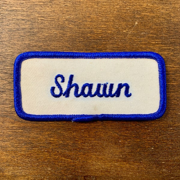 Shawn - A white work shirt name patch that says "Shawn" in blue script with blue border