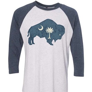 May include: A white and blue baseball tee shirt with a distressed image of a buffalo silhouette with the South Carolina state flag inside. The sleeves are a dark blue color.