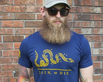 Puede incluir: Camiseta azul marino con un gráfico de serpiente amarilla y el texto "JOIN, or DIE". La gorra de camionero gris tiene un parche azul con una estrella blanca. La persona lleva gafas de sol.