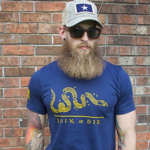 K&ouml;nnte beinhalten: Marineblaues T-Shirt mit gelber Schlangengrafik und dem Text "JOIN, or DIE". Der Mann tr&auml;gt eine graue Trucker-M&uuml;tze mit einem blauen Aufn&auml;her mit einem wei&szlig;en Stern. Der Mann tr&auml;gt eine Sonnenbrille.