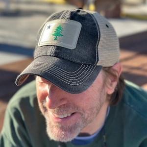 May include: A black and tan trucker hat with a distressed look. The hat features a patch with the state flag of Maine, which depicts a green pine tree and a white star on a blue background.