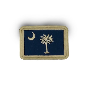 May include: A rectangular patch featuring the South Carolina state flag. The flag has a dark blue background with a white crescent moon in the upper left corner and a white palmetto tree in the center. The patch is bordered with a tan stitching.