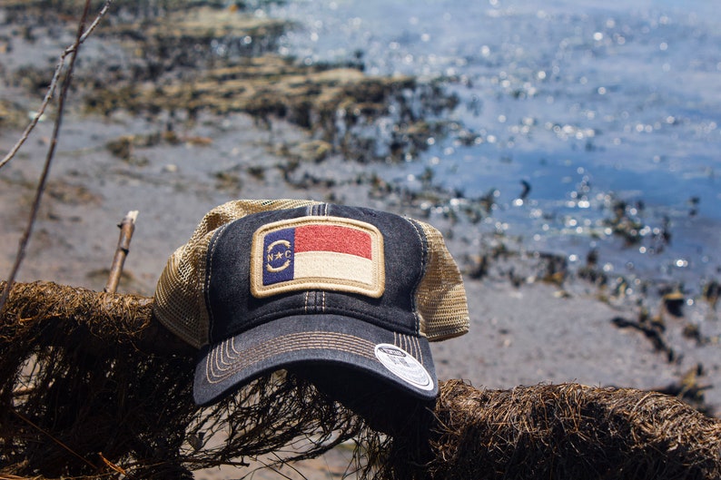 Trucker hat with khaki colored mesh backing, black cotton front panel and bill with tan stitching. Ballcap has an embroidered patch of the North Carolina flag in in the center.