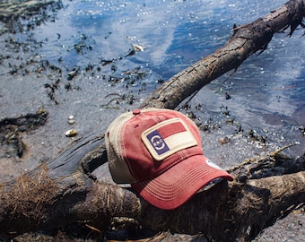 May include: A red and white trucker hat with a North Carolina state flag patch on the front. The hat is sitting on a weathered tree branch.