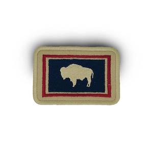 May include: A beige embroidered patch with a red and blue border featuring the state flag of Montana. The flag depicts a white silhouette of a bison on a dark blue background.