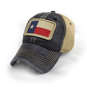 May include: A charcoal gray baseball cap with a tan mesh back and a rectangular patch on the front featuring the Texas state flag. The cap has a curved brim with visible tan stitching and a distressed finish on the dark fabric panels.