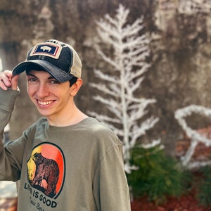 May include: A person wearing an olive green long-sleeve shirt and a baseball cap. The shirt features a bear graphic inside a red and orange circle, with the text "LIFE IS GOOD New Bern". The cap has a bear patch.