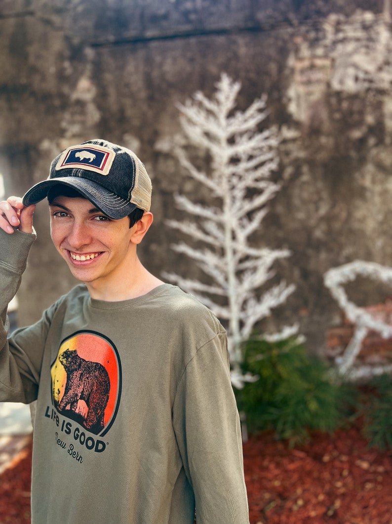 May include: A person wearing an olive green long-sleeve shirt and a baseball cap. The shirt features a bear graphic inside a red and orange circle, with the text "LIFE IS GOOD New Bern". The cap has a bear patch.