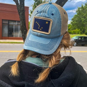 May include: A person wearing a blue and beige trucker hat with a patch featuring the flag of Alaska. The hat has a mesh back and a curved brim.
