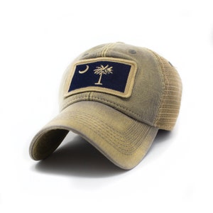 May include: A tan baseball cap with a mesh back and a curved brim. The front features a navy blue patch with a gold crescent moon and a palmetto tree, the state symbol of South Carolina.