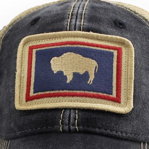 May include: A distressed denim baseball cap with a tan mesh back. The front features a rectangular patch with a navy blue background, a tan bison silhouette, and red and tan borders. The cap has a curved brim and a classic trucker hat design.