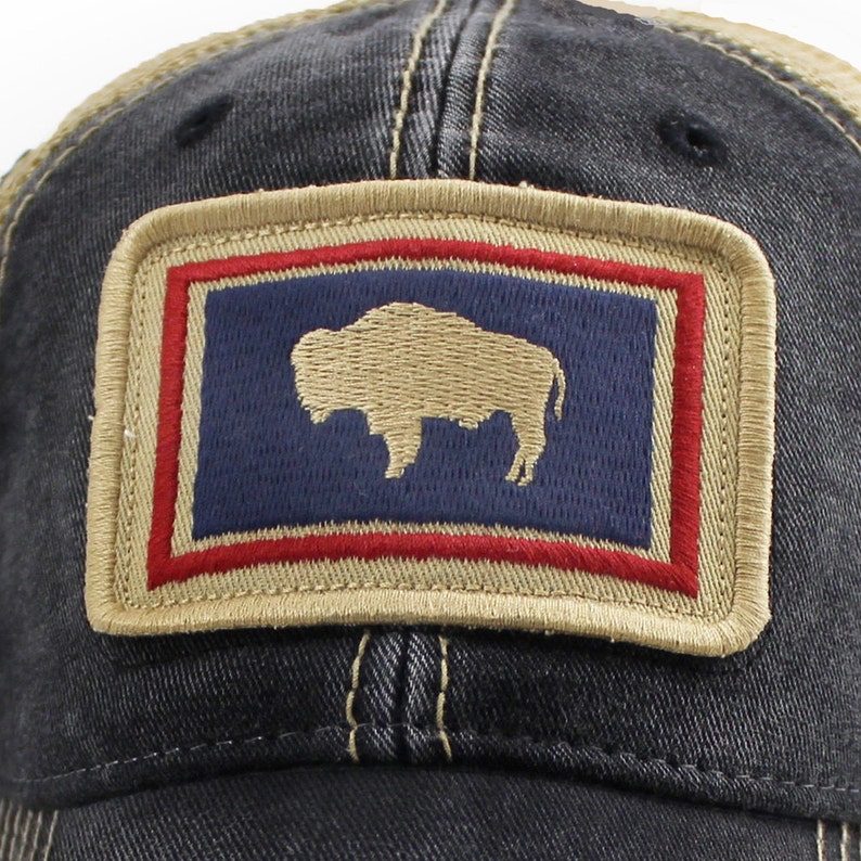 May include: A distressed denim baseball cap with a tan mesh back. The front features a rectangular patch with a navy blue background, a tan bison silhouette, and red and tan borders. The cap has a curved brim and a classic trucker hat design.