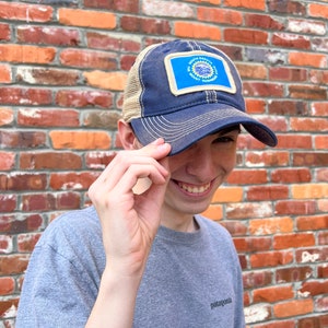May include: A person wearing a blue and tan baseball cap with a patch featuring the South Dakota state seal. The cap has a mesh back and a curved brim. The person is smiling and looking at the camera.