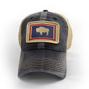 May include: A distressed black trucker hat with a tan mesh back and a curved brim. The front features a rectangular patch with a blue background, a tan bison, and red and tan borders. The hat has a snap closure.