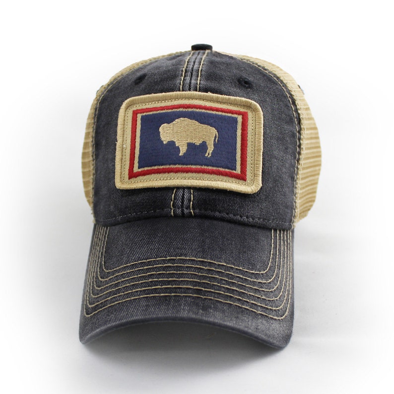 May include: A distressed black trucker hat with a tan mesh back and a curved brim. The front features a rectangular patch with a blue background, a tan bison, and red and tan borders. The hat has a snap closure.