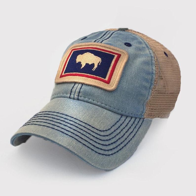 May include: A blue denim baseball cap with a mesh back. The cap features a rectangular patch with a white bison silhouette on a navy blue background, framed by a red and tan border. The cap has a curved brim and a snap closure.