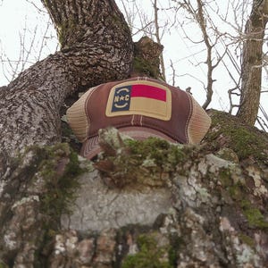 May include: A brown trucker hat with a tan mesh back and a patch featuring the North Carolina state flag and the letters "N*C". The hat is resting on a tree branch.