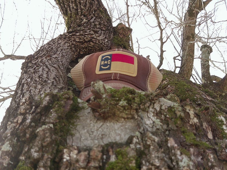 May include: A brown trucker hat with a tan mesh back and a patch featuring the North Carolina state flag and the letters "N*C". The hat is resting on a tree branch.