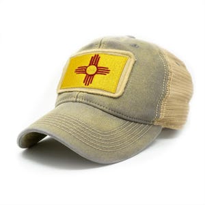 May include: A distressed-look baseball cap with a yellow patch featuring the New Mexico Zia symbol in red. The cap has a khaki-green front and brim, with a tan mesh back. The Zia symbol is a Native American design.