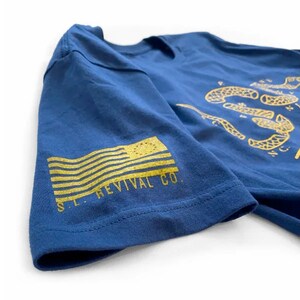 K&ouml;nnte beinhalten: Ein marineblaues T-Shirt mit einer goldenen Schlange und dem Text "NO STEP ON SNEK". Der &Auml;rmel zeigt eine goldene amerikanische Flagge und den Text "S.L. REVIVAL CO."